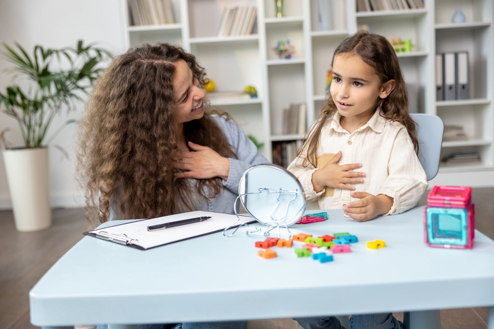 a young brown haired girl sitting at a small therapy table in front of a small mirror with her right hand over her chest, working with her therapist.
