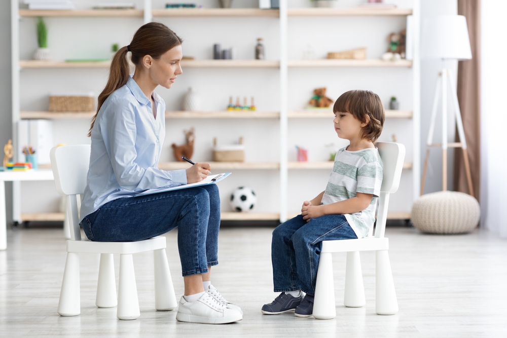 a young boy in a grey striped t-shirt and jeans sitting on a white chair, working directly with a female therapist sitting directly across from him and taking notes.