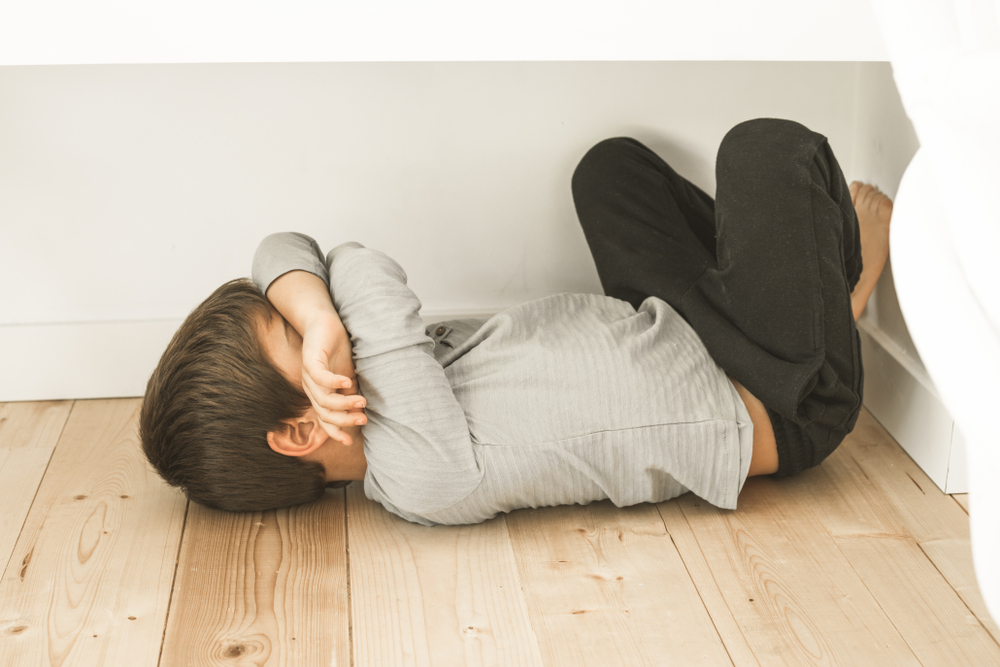 Child lying on the floor in a corner, curled with arms covering their face, in a sparse white room with light wood flooring.