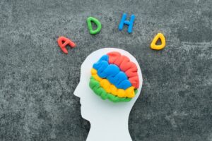 Colorful clay model of a brain inside a white silhouette of a head, with the letters “ADHD” arranged above it on a dark textured background.