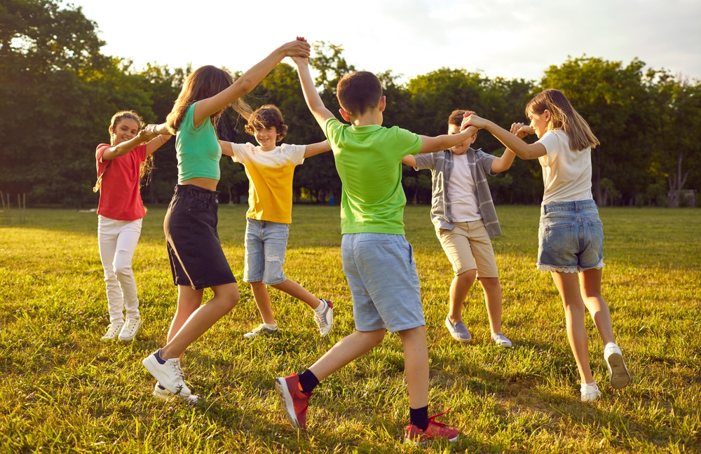 Group of children holding hands and playing together in a grassy field during summer camp, enjoying outdoor activities in a natural setting.