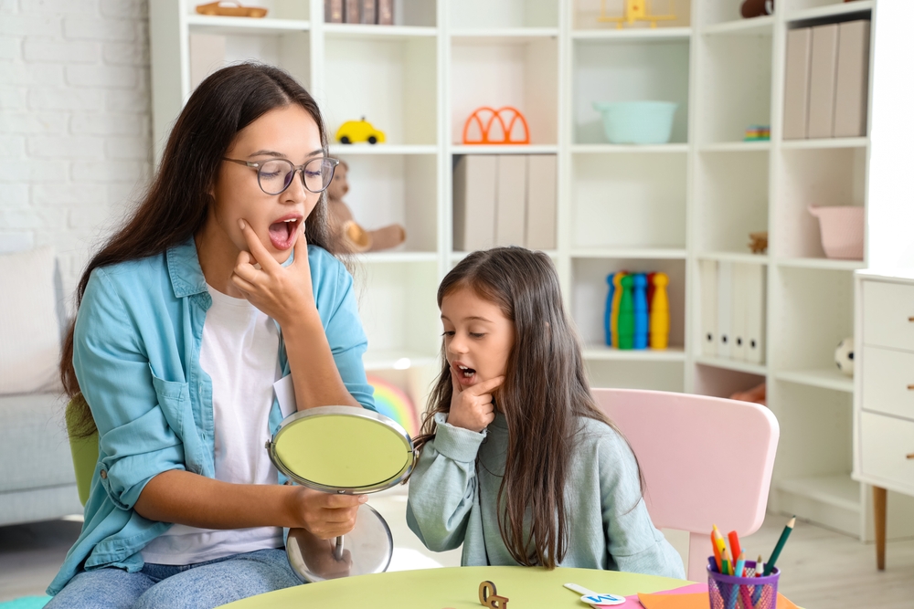 Speech therapist and young girl practicing mouth movements together using a mirror during a speech therapy session.