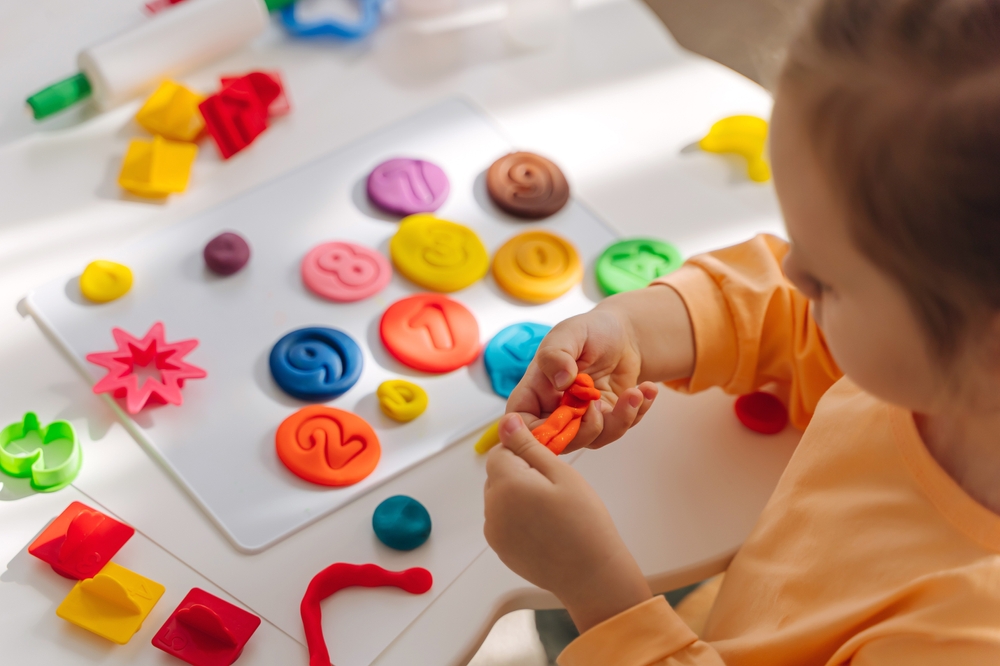 Child playing with colorful clay shapes and numbers on a white table, engaging in a sensory and educational activity.