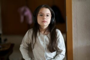 Young girl with long dark hair wearing headphones and glasses, standing indoors with a neutral expression.