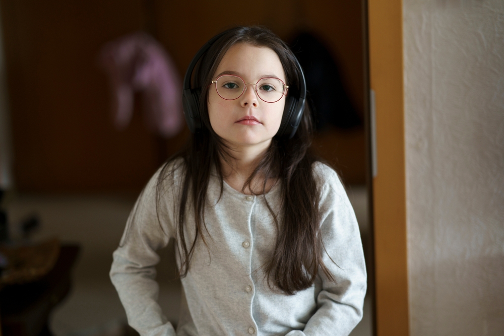 Young girl with long dark hair wearing headphones and glasses, standing indoors with a neutral expression.