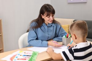 Smiling woman working with a young boy at a table, both drawing with colored pencils, suggesting a supportive and creative learning environment.