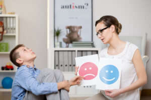 A young boy sits on the floor while a woman holds up two emotion cards—one happy and one sad—helping him identify and express feelings.