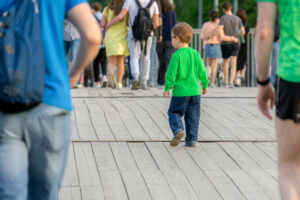 A young boy in a green shirt and jeans walking away on a wooden boardwalk among other pedestrians.