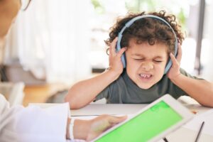 A young child wearing headphones with eyes closed and a distressed expression, covering their ears while an adult shows a tablet in the foreground.