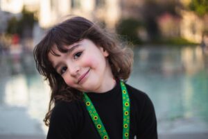 A young child with long brown hair and a gentle smile, wearing a black top and sunflower-patterned lanyard, tilts their head while standing near a reflective pool.