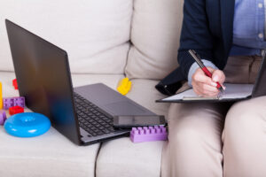 Adult taking notes on a clipboard, sitting next to an open laptop, and toys surrounding the computer.