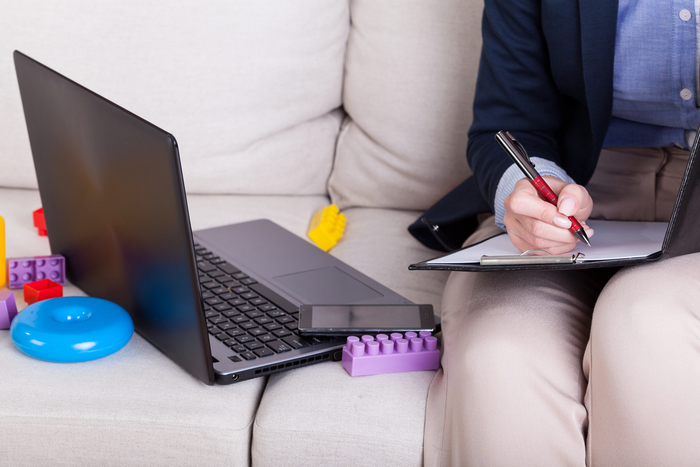 Adult taking notes on a clipboard, sitting next to an open laptop, and toys surrounding the computer.