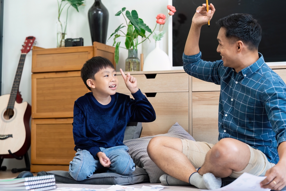 Father and son smiling, both with raised hands, working on a school problem together.