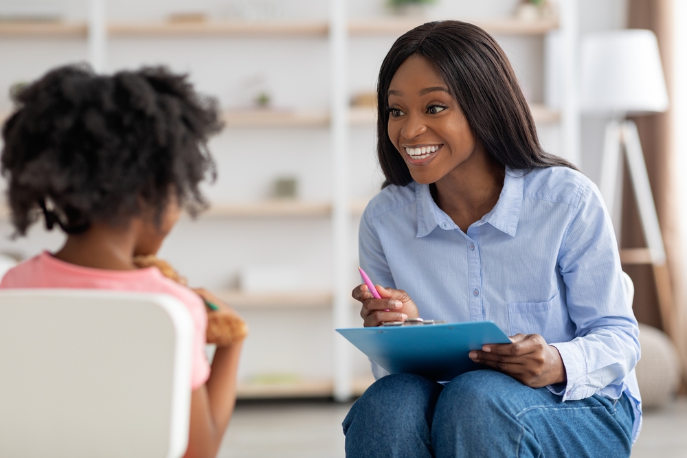 African-American therapist smiling and interacting with a young girl sitting in a chair.