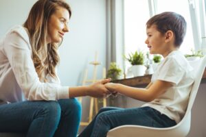 Young boy in a white t-shirt sitting on a chair while a woman with long brown hair wearing a white blouse holds his two hands and looks at him.