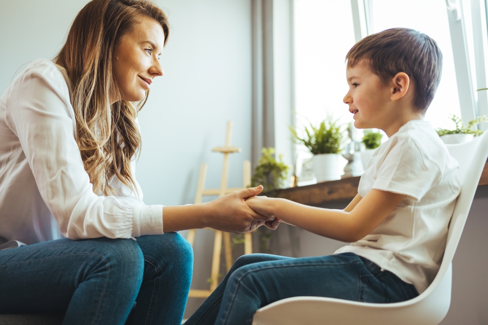 Young boy in a white t-shirt sitting on a chair while a woman with long brown hair wearing a white blouse holds his two hands and looks at him.