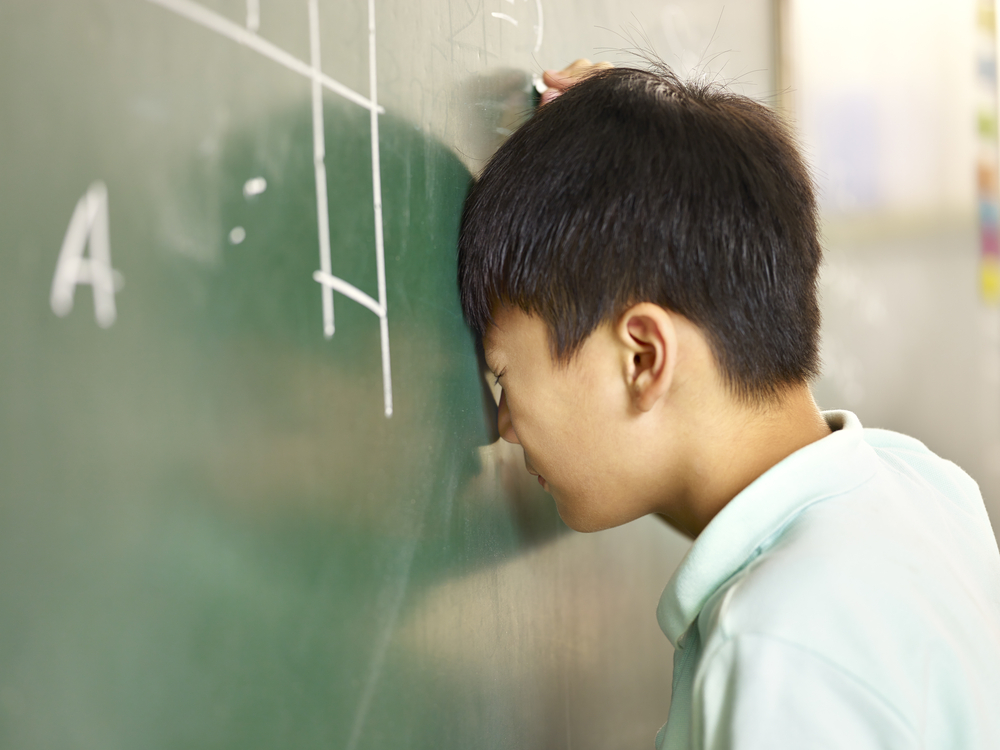 Asian student banging head against green blackboard with chalk in right hand