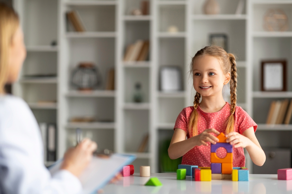 Young girl with braids smiling at the therapist while playing with multi-colored building blocks.