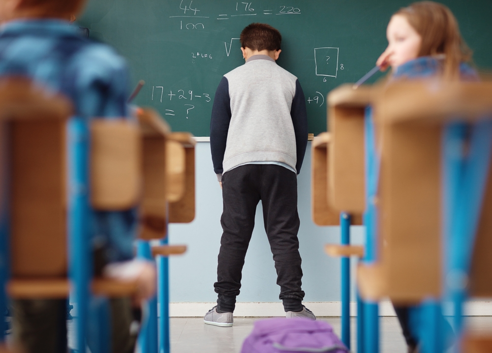Child wearing a football jacket and hands in his pockets leaning his head against a chalkboard.