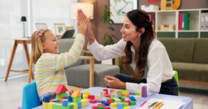 Child and therapist smiling and high-fiving during a play-based therapy session with colorful blocks.