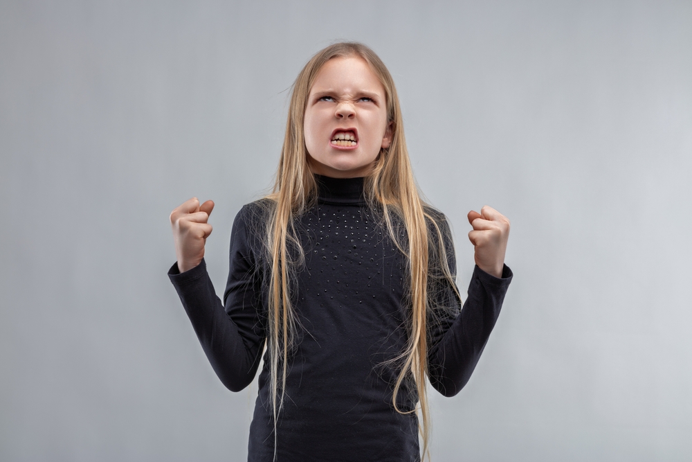  A young girl with long blond hair and a black long-sleeve t-shirt showing her teeth and looking upwards with both fists clenched.
