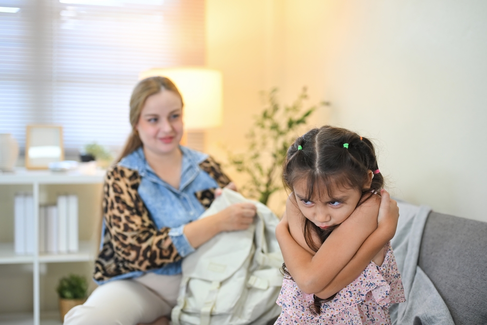 Young brown-haired girl with her arms crossed and her back to an adult holding a school backpack.