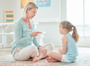 Blond woman in a blue sweater wagging her finger at a young girl seated on a carpet.