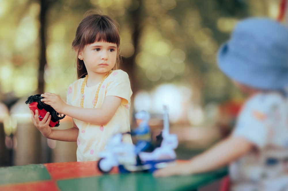 Young brown haired girl in a yellow top taking a toy away from a toddler.
