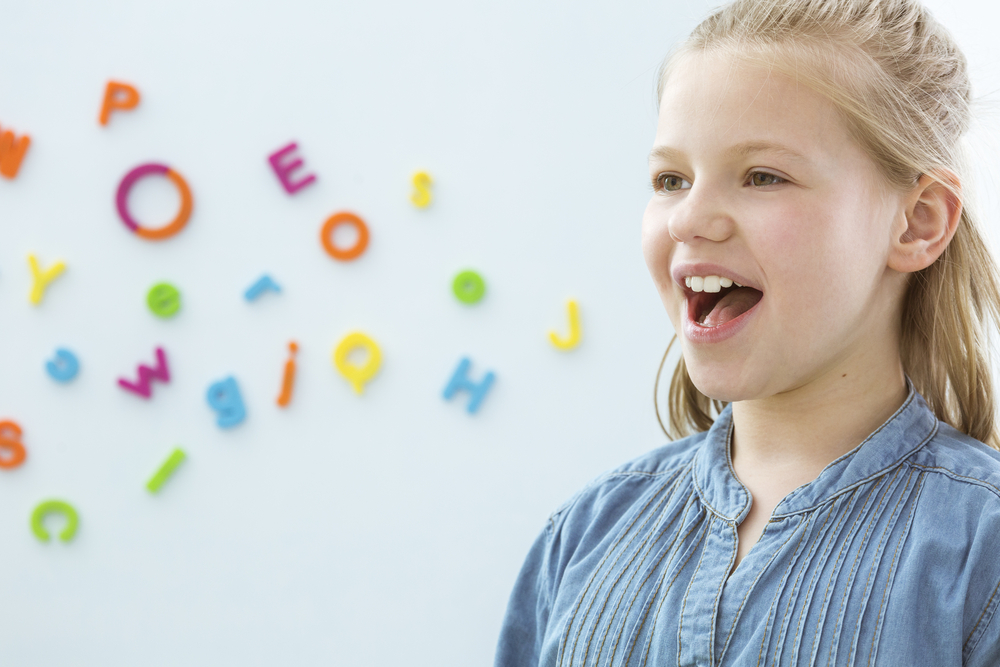 A young girl wearing a denim shirt, and her mouth is open. There are magnetic multicolored letters in the background.