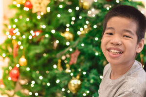 young Asian boy smiling in front of a large, decorated Christmas tree.