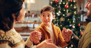 Brown haired boy wearing a yellow cardigan holding his parents' hands near the Christmas tree.