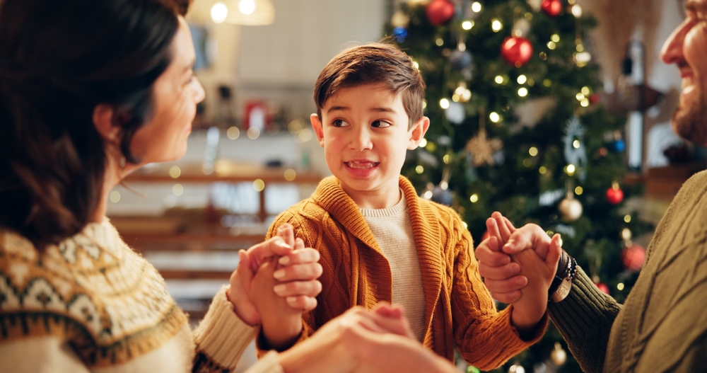 Brown haired boy wearing a yellow cardigan holding his parents' hands near the Christmas tree.