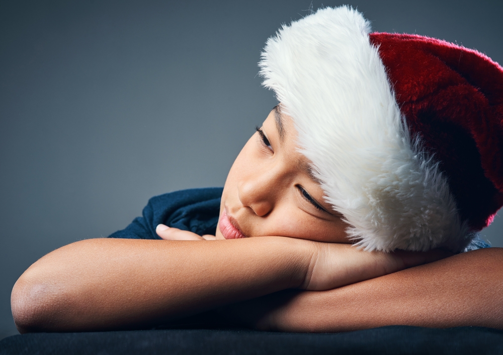 A young Asian boy wearing a Santa hat, resting his head on his arms, looking sleepy.