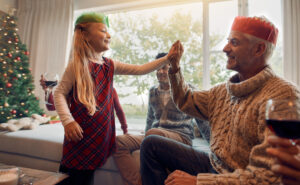 Blond-haired girl giving her grandfather a high-five around the Christmas tree.