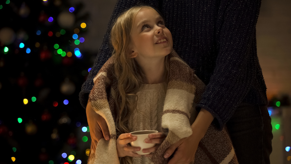 young blond-haired girl holding a hot chocolate wrapped in a brown blanket, looking up at a parent standing by the Christmas tree.