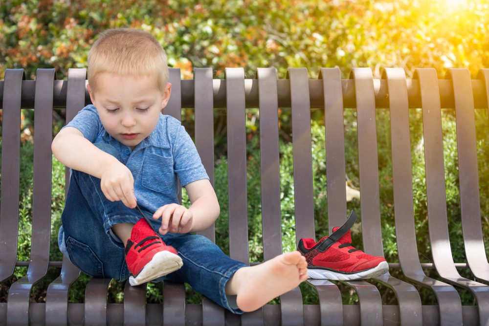 A boy sitting on a black bench, putting on one of his red running shoes.

