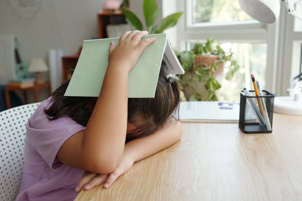 A girl in a pink shirt with brown hair, face down at her desk, and hiding her head under a green book.