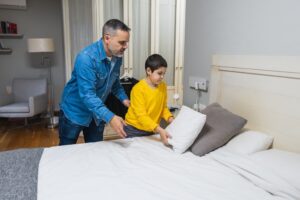 A young boy in a yellow hoodie is learning to make the bed with the help of his father, dressed in a denim shirt.