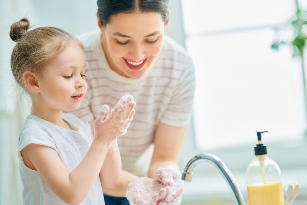 A young girl in a white t-shirt and bun washing her hands with her mom standing over the sink.