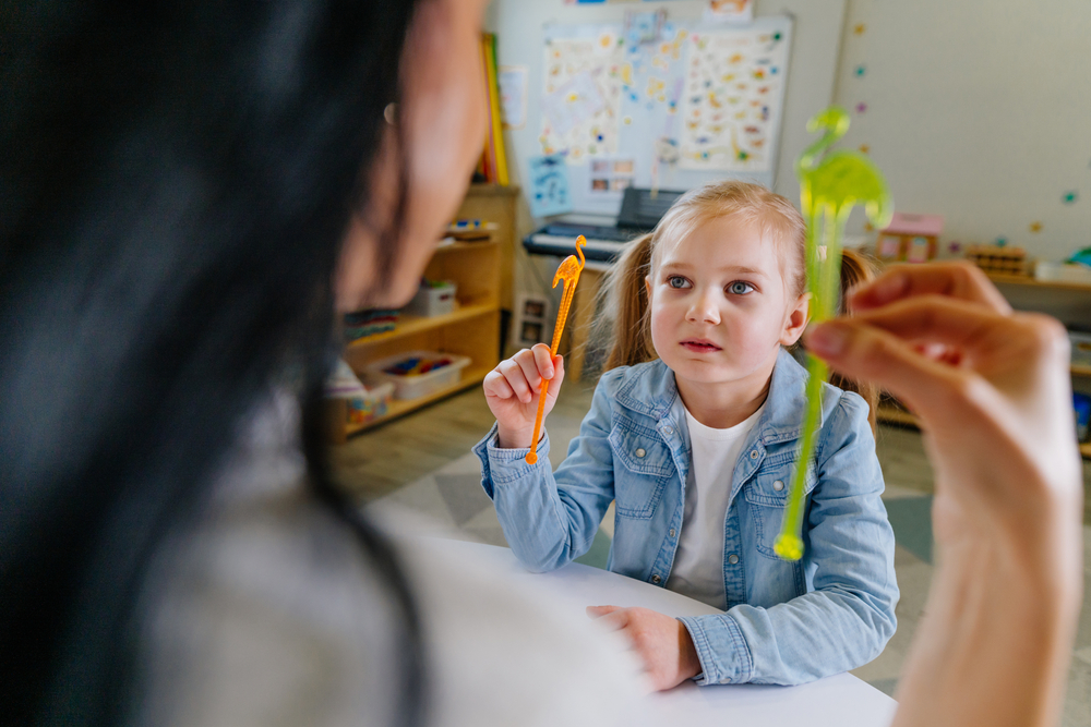 A girl with ponytails wearing a denim jacket holds a plastic flamingo and copies her therapist, who is doing the same.