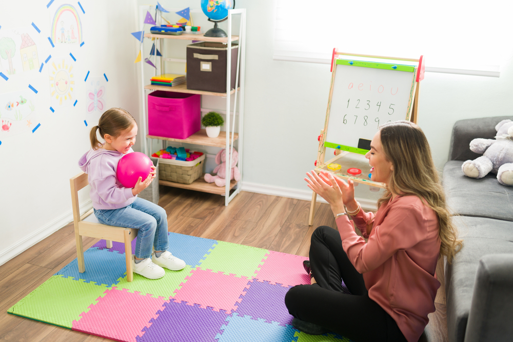 Young girl sitting on a chair holding a pink ball across from her therapist, who is clapping her hands.