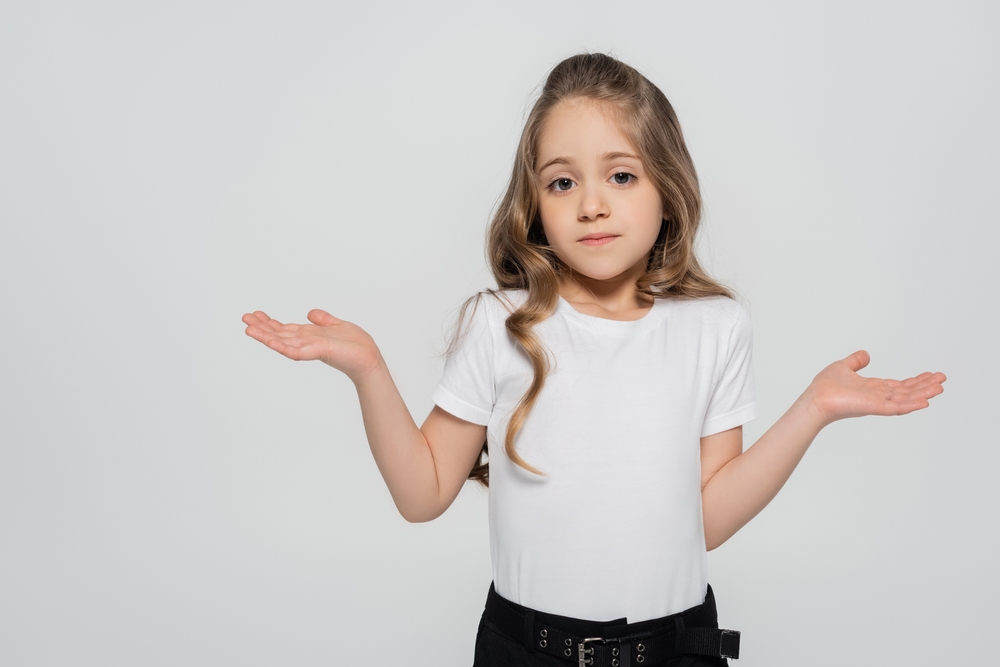 A young brown-haired girl wearing a white shirt, making the confused gesture with her hands because she doesn’t understand.