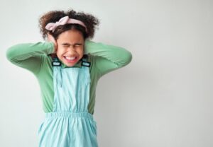 A young African American girl in a green shirt and blue overalls covering her ears and closing her eyes.