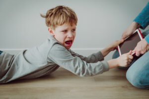 A boy lying on his stomach, tugging his iPad away from his parent while screaming.