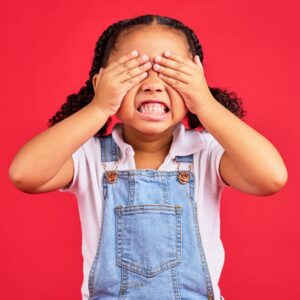 Small African-American girl with ponytails and dressed in denim overalls, clenching her teeth and covering her eyes in frustration.