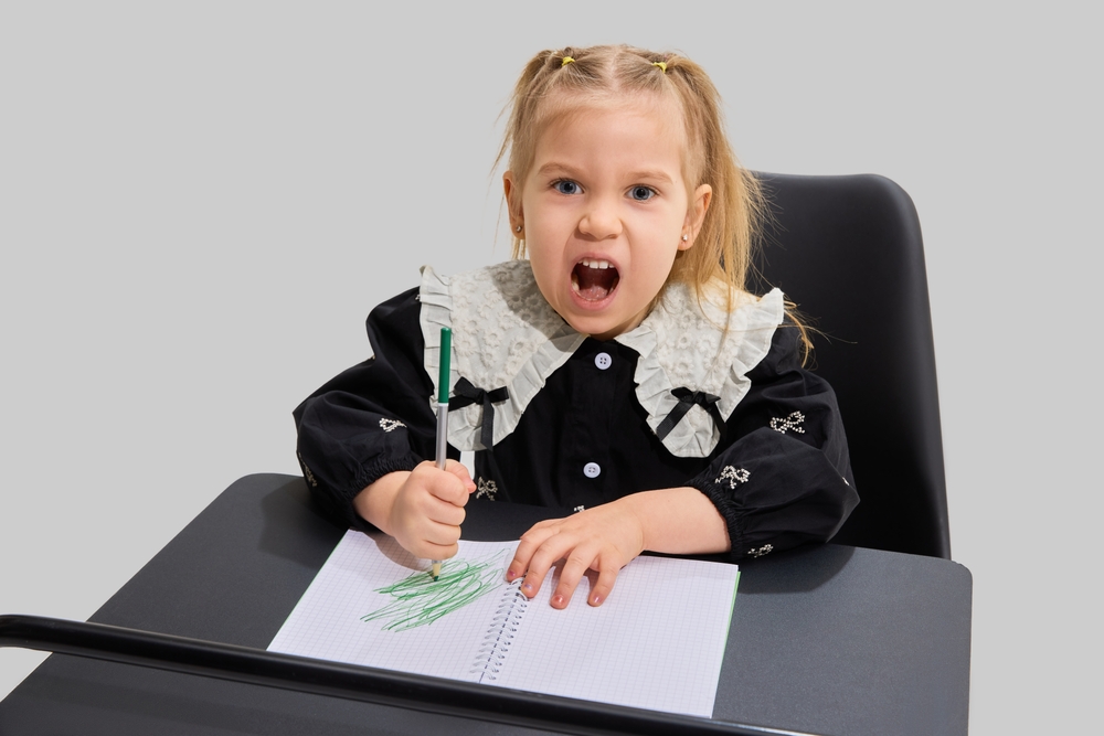 A young blond-haired girl looking frustrated while sitting at a desk and coloring a paper with a green marker.