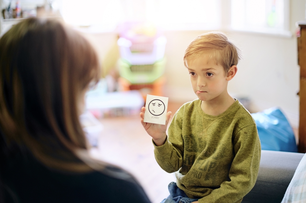 A boy in a green long-sleeved shirt holding a sad face emotion card and showing it to an adult.