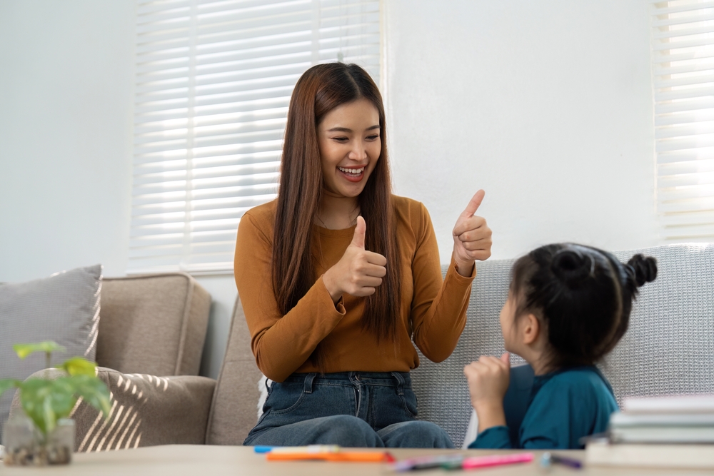 An asian woman in a rust-colored shirt giving two thumbs up to a young girl in a blue shirt.