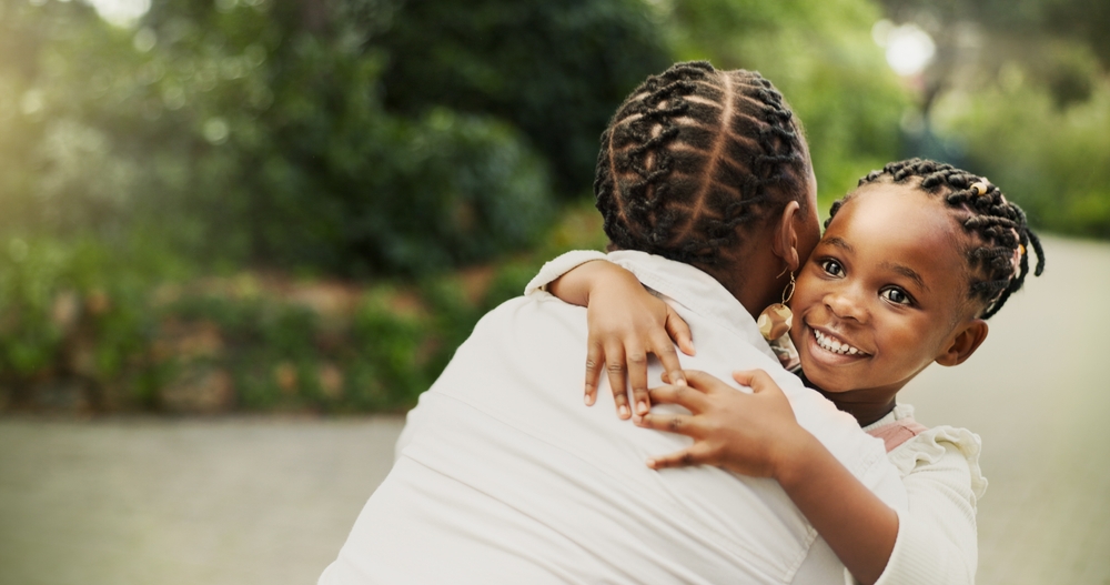 A young African American girl hugging a woman in a white blouse.