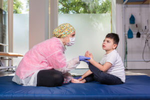 A therapist wearing a face mask, gloves, and a disposable medical gown, working with a severely autistic young boy on a blue mat.